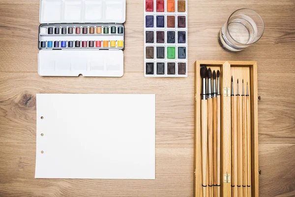 Top view of wooden table with set of paintbrushes, blank paper sheet, water-color and glass of water. Mock up — Stock Photo, Image