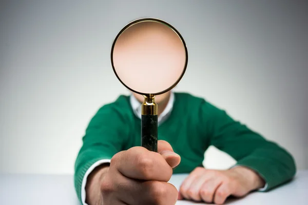 Man in green pullover sitting at white desk and covering face with magnifying glass — Stock Photo, Image