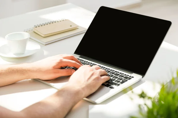 Primer plano del teclado de mano del hombre en el ordenador portátil con pantalla en blanco, colocado en el escritorio blanco con taza de café, bloc de notas y la planta. Prepárate. — Foto de Stock