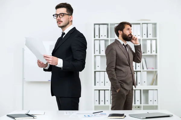 Dois empresários atraentes em pé na mesa do escritório, fazendo papelada e falando ao telefone — Fotografia de Stock
