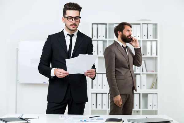 Dois homens de negócios bonitos em pé na mesa do escritório, fazendo papelada e falando ao telefone — Fotografia de Stock