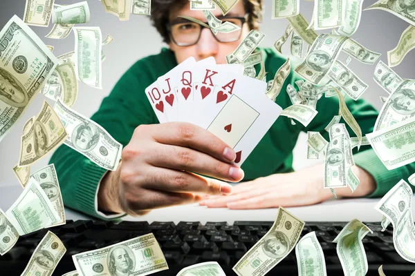 Young man holding royal flash poker cards with keyboard on table and abstract dollar bill rain. Gambling concept