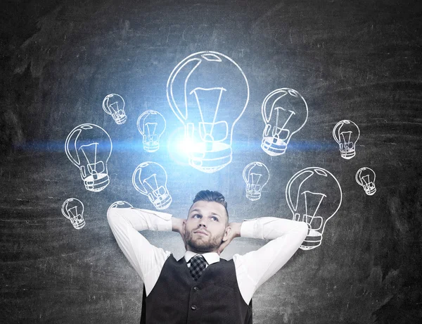 Idea concept with attractive relaxed businessman and illuminated light bulb drawings on chalkboard background — Stock Photo, Image