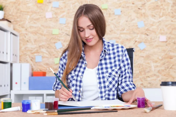 Linda chica con el pelo largo y rubio está pintando en su álbum. Ella está sentada en su lugar de trabajo con mesa, tablero de corcho y estantes de libros. Concepto de boceto para el panorama general —  Fotos de Stock