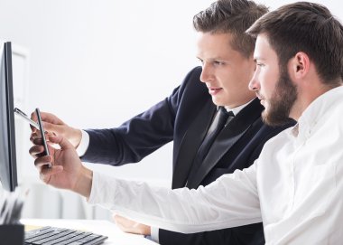 Bilgisayar ve akıllı telefon ile masada iki iş arkadaşı. Birincisi ekranı işaret ediyor, ikincisi de telefonuyla fotoğraf çekiyor. Ortak çalışma kavramı