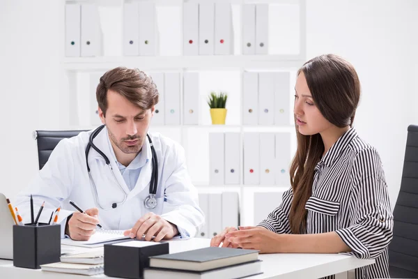 Vrouw in overleg met haar dokter. Hij luistert en maakt aantekeningen om haar te diagnosticeren.. — Stockfoto