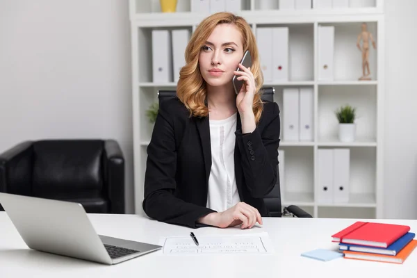 Mulher de negócios lindo está falando em seu telefone celular no escritório. Livros na mesa. Laptop em primeiro plano. Ligantes nas prateleiras. Poltrona de couro preto no canto. Conceito de CEO ocupado no trabalho . — Fotografia de Stock