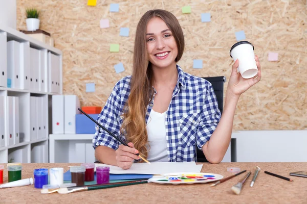 Leende flicka med pensel håller kopp kaffe i handen när du sitter på kontoret med vita bokhyllor. Begreppet konstutbildning — Stockfoto