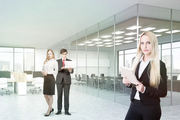 Портрет бізнесменів в офісі. Чоловік і жінка стоять на задньому плані, тримаючи ноутбук. Блондинка на передньому плані з блокнотом. Конференц-зал з бетонними стінами. 3D візуалізація . — стокове фото