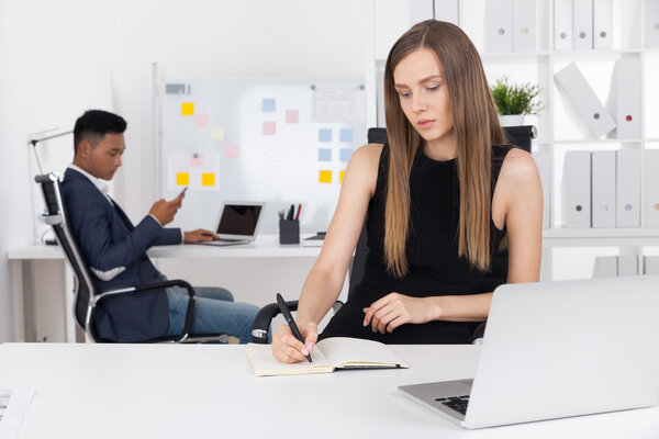 Pretty business lady is taking notes in her notebook. Her Asian colleague is looking at his phone screen. Concept of hard clerk work