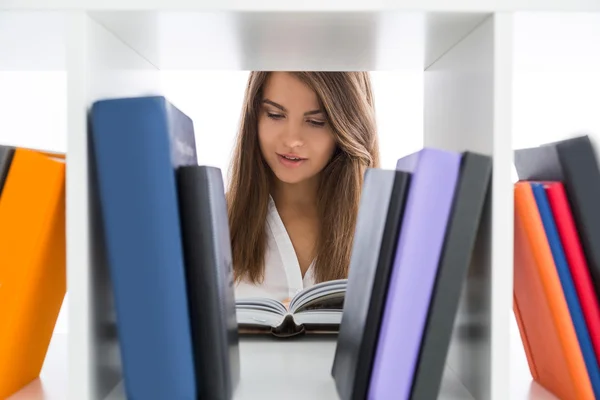 Pretty young lady reading a book standing behind white bookcase. Concept of library and receiving information