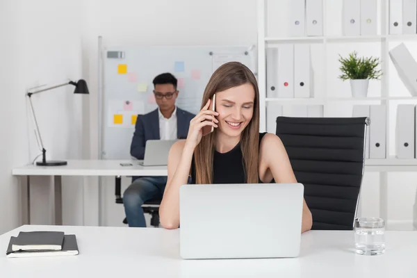 Mosolygó üzleti hölgy fekete ruhában beszél a telefon és a gépelés. A lány koncentrált ázsiai kollégája dolgozik. A menedzseri munka fogalma — Stock Fotó