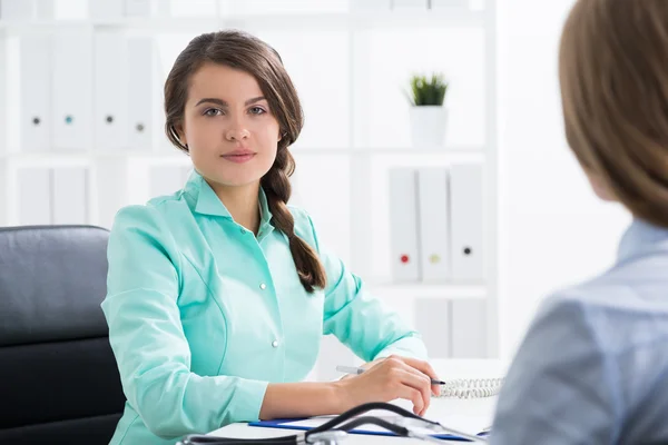 Eine junge Ärztin in grünem Gewand mit geflochtenem Haar spricht mit ihrer Patientin. Konzept des Besuchs beim Kardiologen — Stockfoto