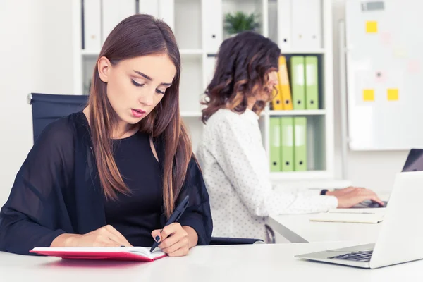 Koncentrerad flicka i svart skriver i sitt anteckningsblock. Hennes kollega är att skriva på laptop tangentbord. Begreppet samarbete — Stockfoto