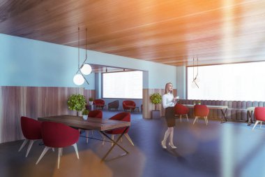 Genç bir kadın, mavi ve ahşap duvarları, beton zemini ve kırmızı koltuklu yemek masalarıyla modern yemek odasında yürüyor. Tonu ayarlanmış resim