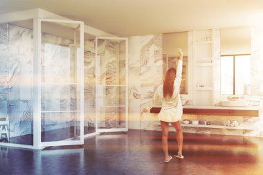 Beyaz mermer duvarları, gri zemini, duş kabini ve çift lavabosu olan lüks bir banyoda duran genç kadının arka manzarası. Tonu ayarlanmış resim