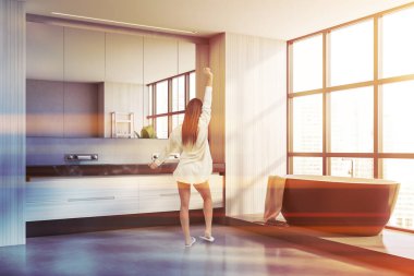 Modern panoramik banyoda beyaz duvarları, beton zemini, rahat bir küveti ve çift lavabosu olan genç bir kadının dikiz görüntüsü. Tonu ayarlanmış resim