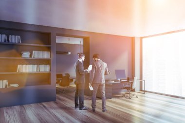 Portrait of two businessmen talking in CEO office corner with gray walls, wooden floor, computer table and bookcase. Toned image