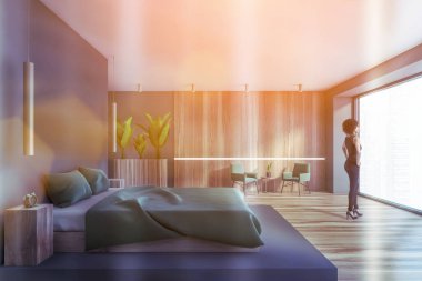Young African American woman standing in modern bedroom with blue and wooden walls, wooden floor and comfortable king size bed. Toned image