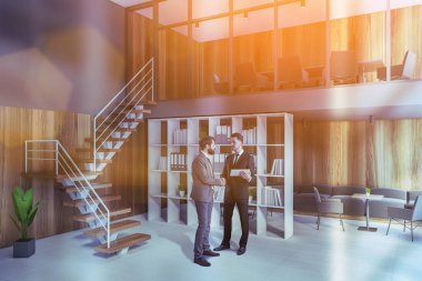 Portrait of two businessmen talking in modern open space office with blue and wooden walls, concrete floor and waiting room in background. Toned image