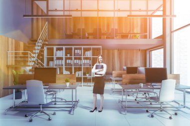 Businesswoman standing in modern open space office with blue and wooden walls, concrete floor and rows of computer tables. Toned image
