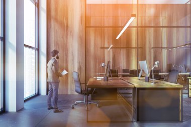 Young businessman with documents standing in modern CEO office with wooden walls, concrete floor and computer table. Toned image