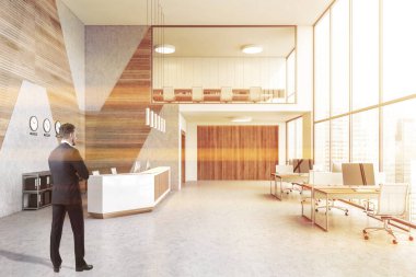 Young businessman standing in moder office with white and wooden walls, concrete floor, row of computer tables and reception desk. Toned image
