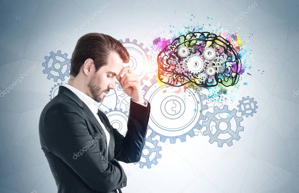 Side view of pensive young businessman standing near gray wall with with colorful brain sketch drawn on it. Concept of brainstorming.