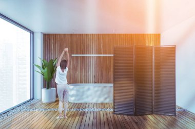 Modern banyoda beyaz ve ahşap duvarları, ahşap zemini ve beyaz küveti olan genç bir kadının dikiz görüntüsü. Tonu ayarlanmış resim