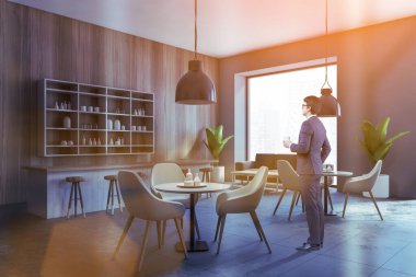 Genç işadamı, ahşap duvarları, beton zemini ve yuvarlak masaları olan modern bir kafede duruyor. Tonu ayarlanmış resim