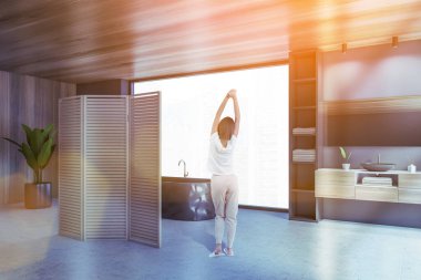 Modern banyo köşesinde beton ve ahşap duvarları, beton zemini, lavabosu ve siyah küveti olan genç bir kadın. Tonu ayarlanmış resim