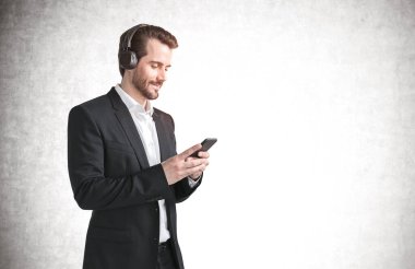 Beton duvarın yanında akıllı telefon ve kulaklık kullanan gülümseyen genç iş adamının portresi. İletişim kavramı. Model