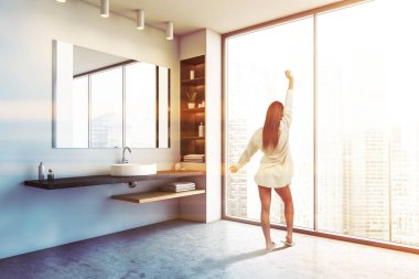 Panoramik banyoda beyaz duvarları, beton zemini olan ve büyük bir aynayla batan genç bir kadının dikiz görüntüsü. Tonu ayarlanmış resim