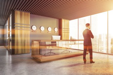 Gri ve ahşap duvarları ve saatli resepsiyon masasıyla modern ofis salonunda duran genç işadamının arka manzarası. Tonu ayarlanmış resim