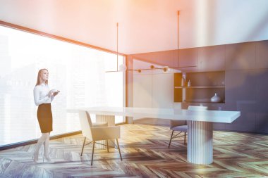 Gri duvarları, ahşap zemini ve beyaz masası olan modern yemek odasında duran genç ve güzel bir kadın. Tonu ayarlanmış resim