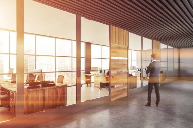 Gri ve ahşap duvarları ve arka planda açık uzay ofisiyle modern ofis salonunda duran genç işadamı. Tonu ayarlanmış resim