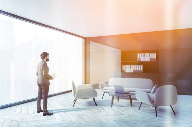 Gri duvarları, ahşap zemini ve rahat beyaz koltuğu olan panoramik oturma odasında duran genç adam. Tonu ayarlanmış resim