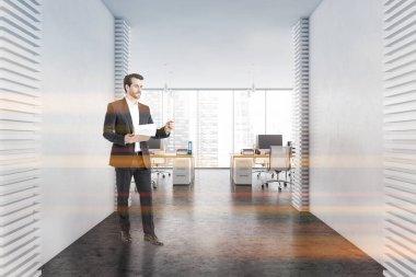 Genç iş adamı, beyaz duvarları, beton zemini ve beyaz bilgisayar masalarıyla modern açık uzay ofisinde duruyor. Tonu ayarlanmış resim