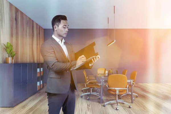 年轻的亚洲商人 有文件夹 站在现代开放的办公室 有灰色和木制的墙壁 木制的地板和电脑桌 被投掷的图像 — 图库照片