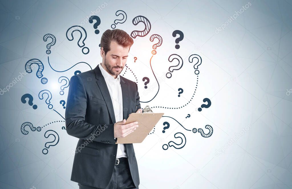 Portrait of bearded young businessman with clipboard standing near gray wall with question marks drawn on it. Mock up