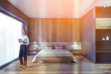 Genç bir kadın, gri ve ahşap duvarları, ahşap zemini ve rahat kral yatağı olan modern ana yatak odasında duruyor. Tonu ayarlanmış resim