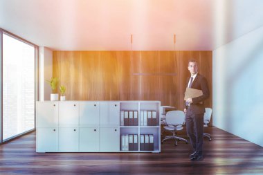 Modern açık uzay ofisinde beyaz ve ahşap duvarları, ahşap zemini ve bilgisayar masaları olan genç bir işadamı. Tonu ayarlanmış resim