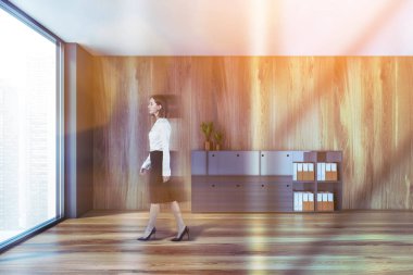 Gri ve ahşap duvarları, ahşap zemini ve gri dolabı olan modern ofis bekleme odasında yürüyen bulanık genç bir kadın. Tonu ayarlanmış resim