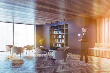 Modern oturma odasında ya da kütüphane köşesinde gri duvarları, ahşap zemini, kitaplığı ve rahat bej koltukları olan güzel Avrupalı bir kadın kahve içiyor. Tonu ayarlanmış resim