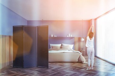 Modern yatak odasında, gri ve ahşap duvarları, ahşap zemini ve rahat kral yatağı olan genç bir kadının dikiz görüntüsü. Bulanık şehir manzaralı bir pencere. Tonu ayarlanmış resim