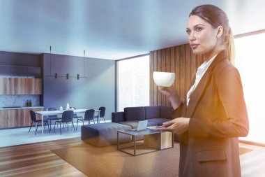 Genç bir kadın, gri oturma odasının köşesinde, tahta zemin ve rahat bir kanepe ile kahve içiyor. Mutfak arka planda. Tonu ayarlanmış resim