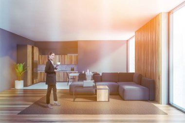 Gri oturma odasında, tahta zeminli ve rahat kanepeli kahve içen genç bir kadın. Mutfak arka planda. Tonu ayarlanmış resim