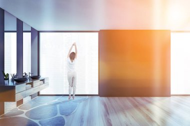 Modern banyoda, gri duvarları, çift lavabosu ve bulanık manzaralı pencereleri olan genç bir kadının dikiz görüntüsü. Tonu ayarlanmış resim