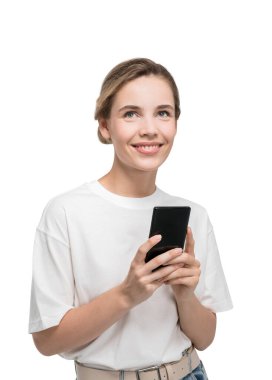 Beyaz arka planda izole edilmiş çekici genç bir kadın elinde akıllı bir telefonla gülümsüyor ve hoş şeyler hayal ediyor.