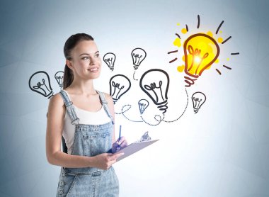 Kot pantolon giymiş Avrupalı çekici genç bir kadın. Üzerinde renkli iş lambası çizimi olan bir önlük giyiyor ve duvarın yanında kafa yoruyor. Yaratıcılık ve beyin fırtınası kavramı